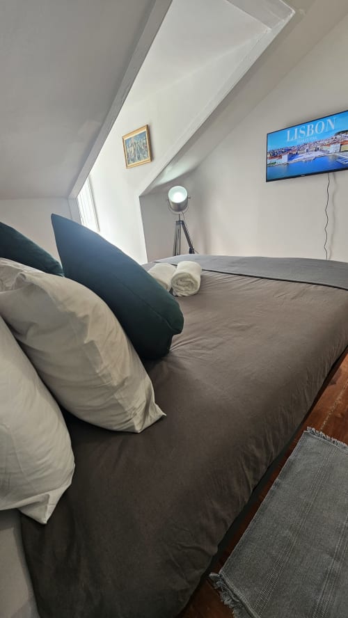 Quarto aconchegante de albergue em Lisboa com cama, almofadas, cabeceira escura, TV de tela plana exibindo paisagem urbana de Lisboa, luminária tripé e