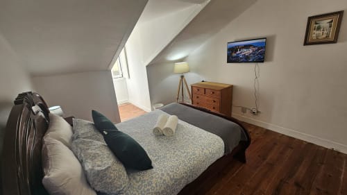 Quarto aconchegante de hostel em Lisboa com cama de casal, cômoda de madeira, TV tela plana e vista da cidade pelo teto inclinado