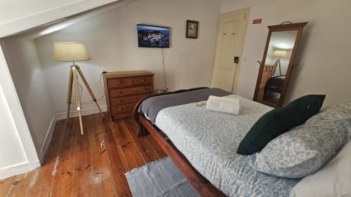 Quarto luminoso de albergue em Lisboa com duas camas, cômoda de madeira, luminária em tripé e vista da cidade na TV