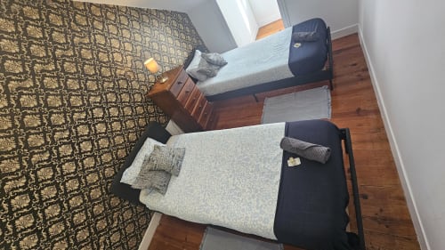 Quarto de hostel em Lisboa com duas camas individuais, móveis de madeira, carpete estampada e vista para a cidade pela janela