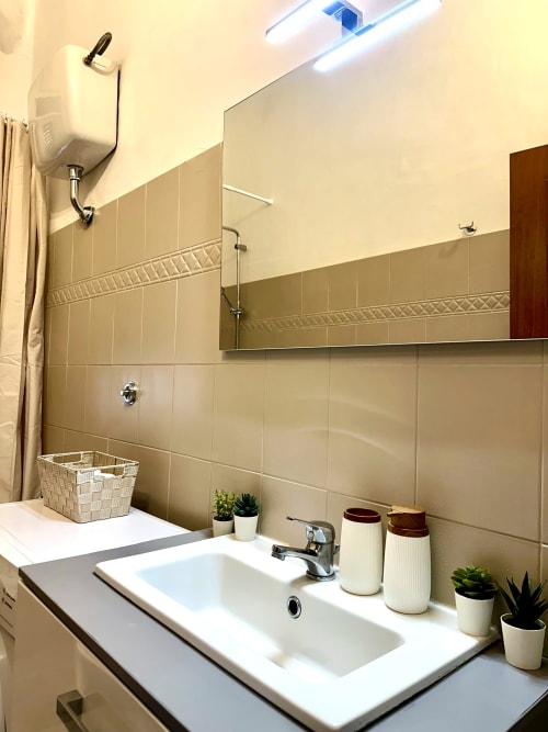 Nowoczesna łazienka z białą umywalką, beżowymi płytkami, lustrem i roślinami doniczkowymi w apartamencie RomeAntique Stay w Rzymie