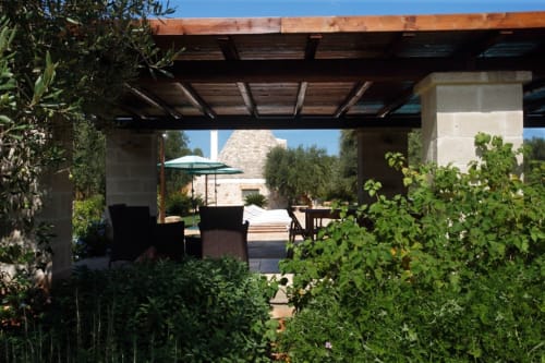 Terrazza Trulli Boccadoro con pergolato, area salotto, ombrellone blu e giardino verdeggiante sotto cielo sereno