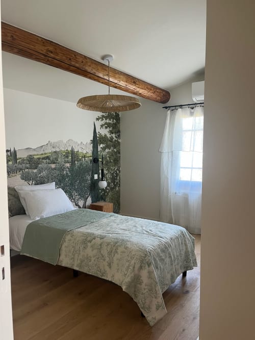 Chambre de la villa Soleù avec poutre en bois apparent, luminaire suspendu tissé et tête de lit murale paysage méditerranéen