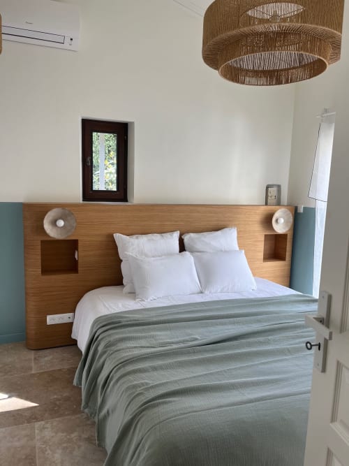 Chambre moderne de la villa Soleù avec tête de lit bois, oreillers blancs, plaid rayé vert, luminaire suspendu tissé et vue sur le jardin