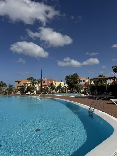 Piscina de aguas cristalinas en el complejo Kamalahouse de Corralejo, rodeada de coloridos edificios y palmeras bajo un cielo despejado