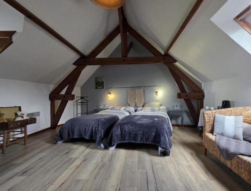 Spacious attic bedroom with exposed wooden beams, twin beds, wicker chairs, and hardwood floors at Ferme de la Forterie