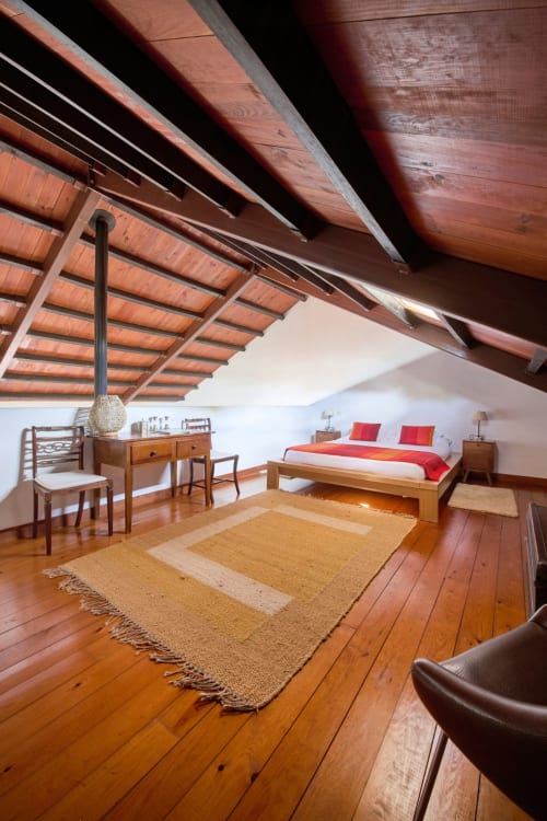 Quarto no sótão da Quinta da Alcaidaria-Mór com vigas de madeira aparentes, cama queen com detalhes em vermelho, móveis de madeira e tecidos artesanais