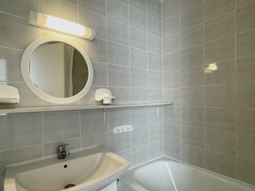 Salle de bain moderne avec miroir rond, lavabo, baignoire et murs carrelés gris à l'appartement Le Vallon des Sources