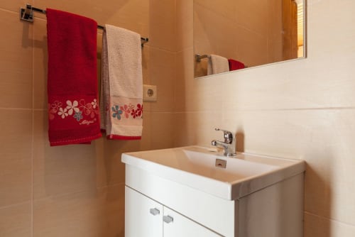 Banheiro moderno com pia branca, torneira cromada, toalhas decorativas vermelhas e bege no suporte de parede, e paredes revestidas de azulejos bege