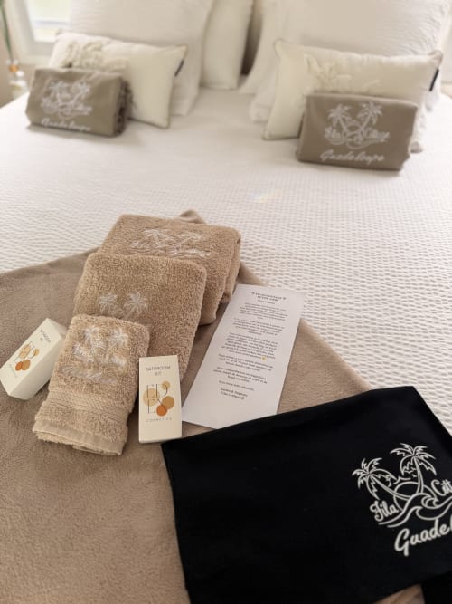 Filao Cottage welcome amenities on bed: towels with embroidered palm design, cosmetics, information leaflet, and black tote