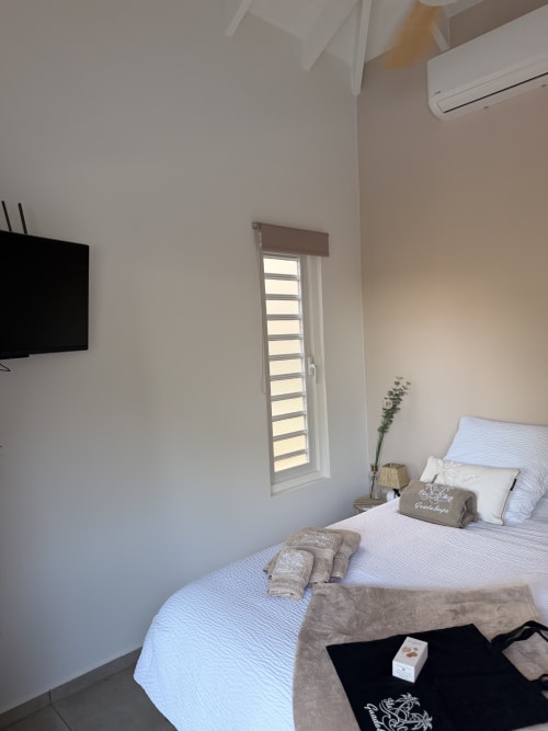Modern bedroom at Filao Cottage in Sainte-Rose with wall-mounted TV, louvered window, and white bedding