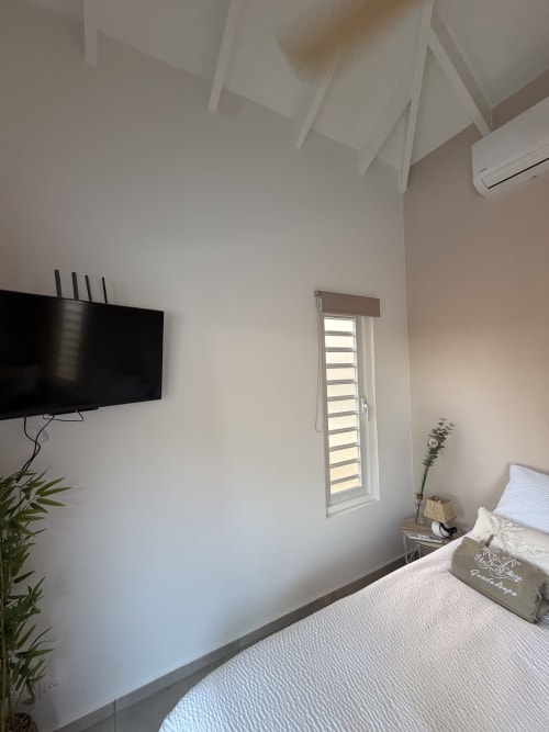 Modern bedroom at Filao Cottage with wall-mounted TV, air conditioning unit, louvered window, and white bedding in