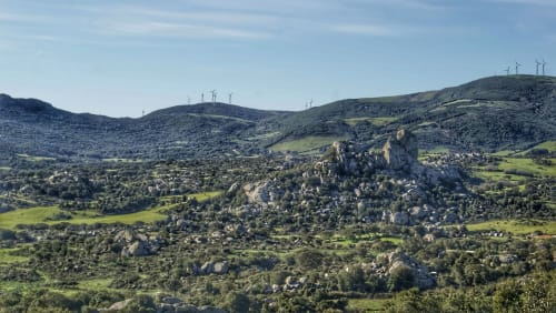 Lussureggiante valle verde con massi sparsi e vegetazione vicino a Valledoria, Italia, con turbine eoliche sulle colline lontane