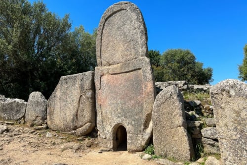 Antica struttura megalitica in pietra con ingresso ad arco presso Valledoria, Italia, circondata da massi e paesaggio mediterraneo