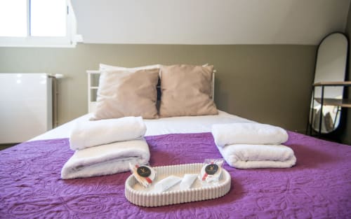 Bright bedroom at La Conciergerie Millau apartment with purple quilted bedspread, folded towels, and welcome amenities on bed
