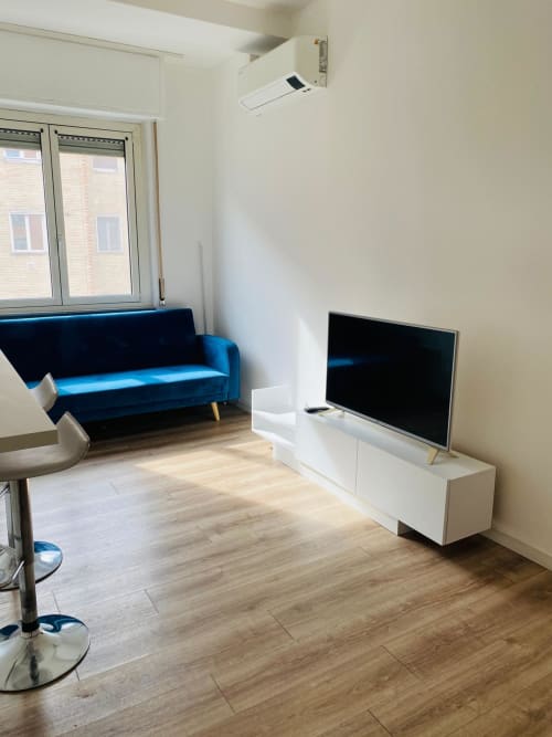 Modern living room in Milano Underground apartment with blue velvet sofa, flat-screen TV on white stand, wooden floors, and