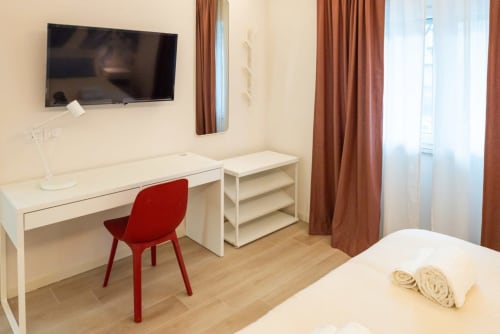 Camera Deluxe Suite Flavia presso EQM Roma con scrivania, sedia rossa, TV e finestre con vista sulla città con tende arancioni