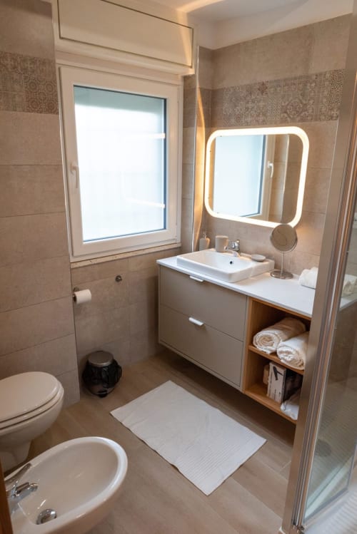 Bagno moderno nella Suite Ottavia Deluxe dell'EQM Roma con specchio illuminato, lavabo bianco, WC, bidet e doccia