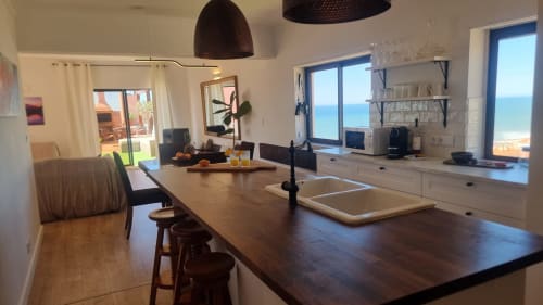 Cozinha e área de refeições moderna em apartamento em Ericeira com vista para o mar através de grandes janelas, bancada escura e mesa de madeira