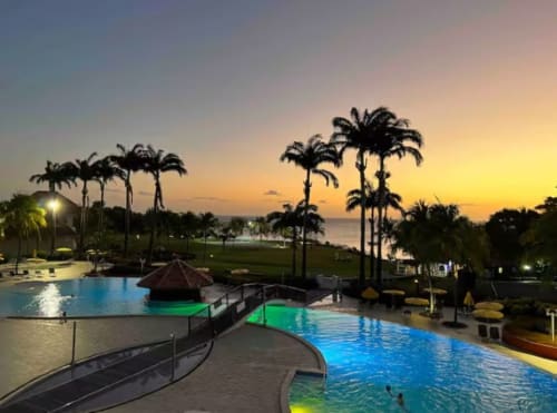 Espace piscine du Tropic Appartements au coucher de soleil avec palmiers, bassins illuminés et vue sur mer à Sainte-Anne, Martinique