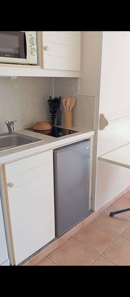 Kitchenette compacte avec armoires blanches, plaque de cuisson, réfrigérateur et ustensiles de cuisine aux Tropic Appartements