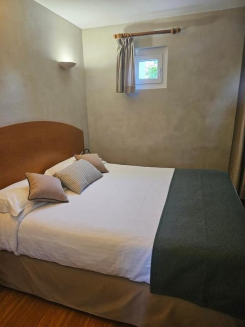 Chambre double confortable avec lit en bois, linge blanc, oreillers rayés, applique murale, petite fenêtre avec rideaux
