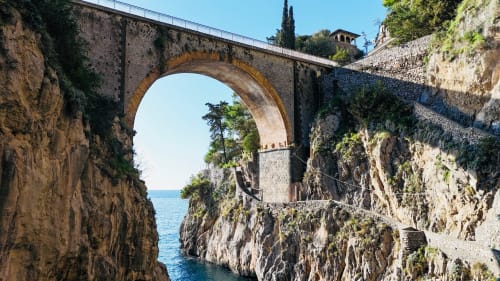 Antico ponte in pietra con arco che attraversa scogliere rocciose costiere e mare turchese a L'Amaranto, Agerola Napoli