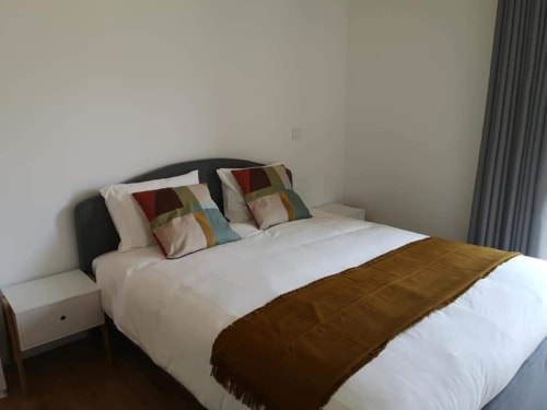 Quarto espaçoso com roupas de cama brancas, almofadas coloridas, manta marrom, cabeceira escura e cortinas cinzentas na Casa da Avó Ana