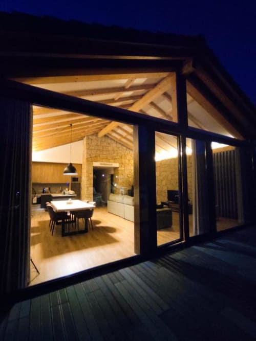 Moderno interior de villa à noite com área de jantar iluminada, vigas de madeira e grandes portas de vidro abertas para o terraço, Mondim