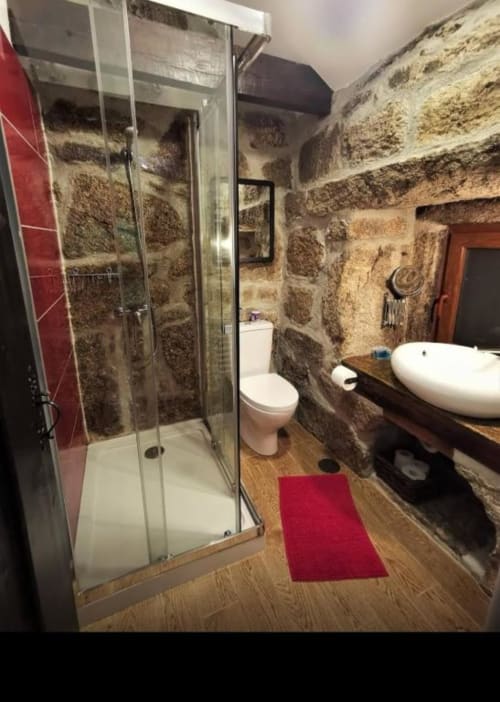 Banheiro rústico de pedra com box de vidro, pia branca, vaso sanitário e piso de madeira na vila Casa da Avó Ana