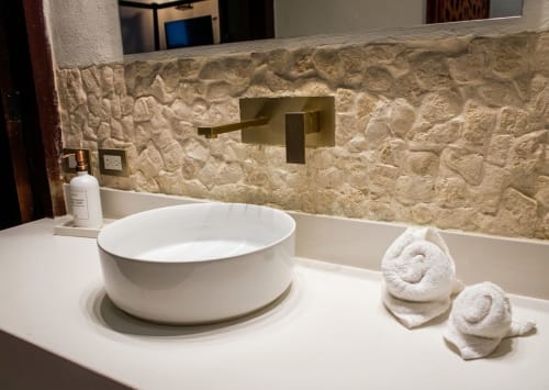 Baño de lujo en Villa Merida Boutique Hotel con lavabo blanco, backsplash de piedra texturizada, grifo de latón y toallas enrolladas