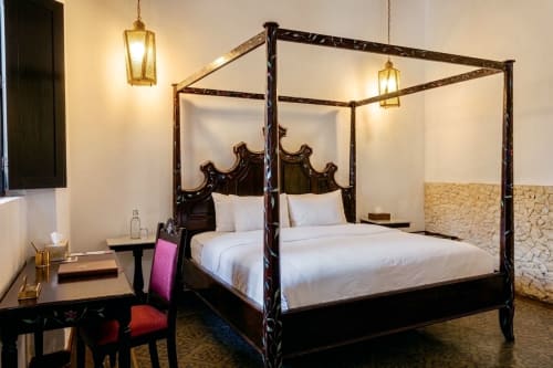 Elegante cama dosel en Villa Merida Boutique Hotel con marco de madera oscura ornamentada, ropa blanca y iluminación colgante dorada