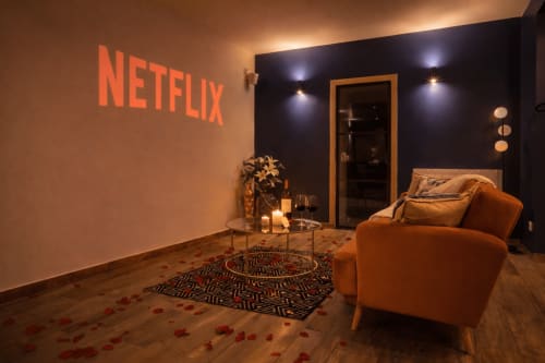Salon Netflix au SPA LE CLANDESTIN Cambrai avec fauteuil orange, écran de projection et pétales de roses romantiques