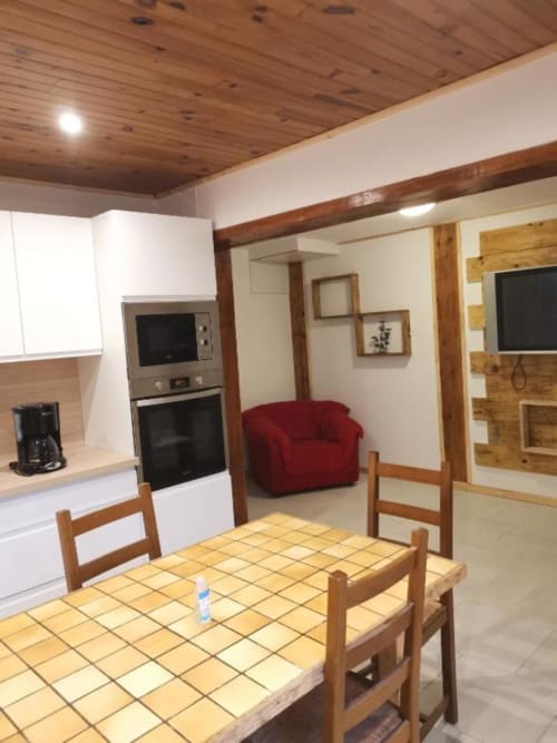 Cottage kitchen-dining area with wooden table, beamed ceiling, white cabinetry, and view into living room with red armchair,