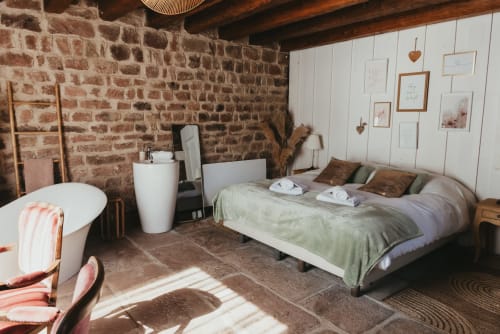 Rustic bedroom cottage with exposed brick walls, wooden beams, bed with green bedding, and white modern bathroom fixtures at