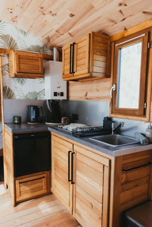 Wooden cabin kitchen in La Roulotte cottage at Les Cabanes du Nideck with wooden cabinets, dark countertop, and window view,