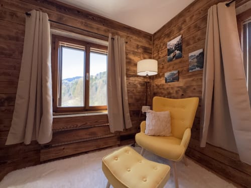 Cozy mountain chalet reading nook with yellow armchair, wood walls, and panoramic alpine views through large windows in Val