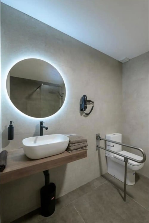 Baño moderno en ML Apartments Valencia con espejo circular iluminado, lavabo blanco y barras de apoyo accesibles