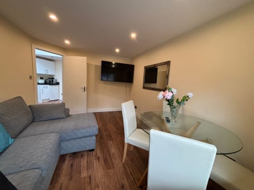 Modern living room with gray sofa, dining table, wall-mounted TV, and kitchenette in England Furnished Homes villa, High