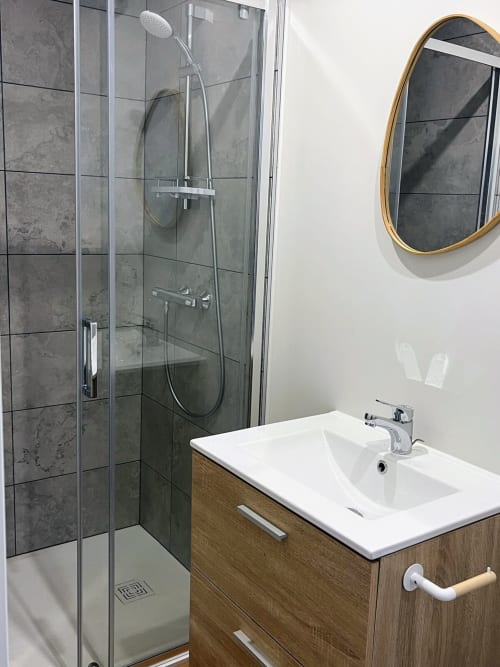 Salle de bain moderne avec douche vitrée, lavabo blanc, meuble de rangement en bois et miroir à cadre doré aux Logis du canal à Betton