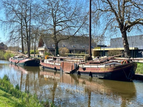 Péniches historiques amarrées sur le canal aux Logis du canal, Betton. Propriété riveraine avec arbres nus et bâtiments traditionnels.