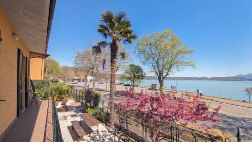 Villa italiana in riva al lago con palme, giardino fiorito rosa, terrazza con balcone che affaccia su acqua e montagne