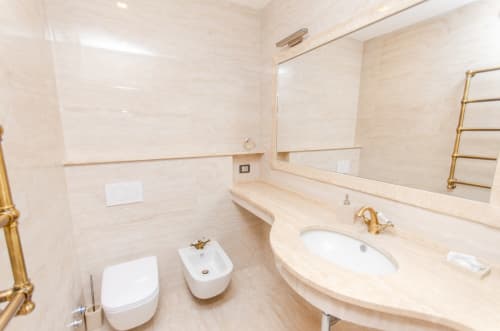 Bagno moderno con piastrelle di marmo beige, vanity con lavabo curvo, WC, bidet, rubinetteria dorata e scaldasalviette a Milano