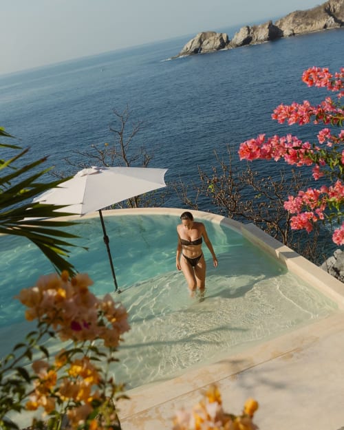 Mujer en bikini en piscina infinita con vista a la bahía de Puerto Ángel, acantilados rocosos, buganvilias rosas y blanco