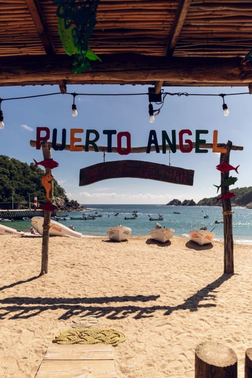 Entrada de la playa Puerto Ángel con cartel colorido, botes y agua turquesa en el resort Casa Baori en México