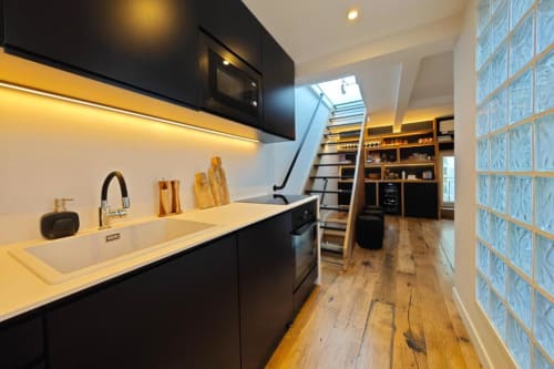 Cuisine moderne de l'appartement O LIT avec meubles noirs, plan de travail blanc, parquet et détail de mur en brique de verre bleu
