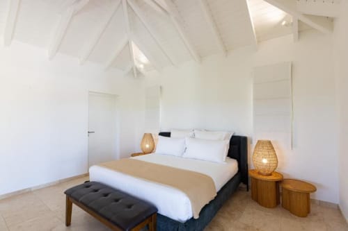 Chambre lumineuse et moderne avec plafond voûté blanc, lit king-size, lampes suspendues tressées, tables de nuit en bois et vue sur l'océan