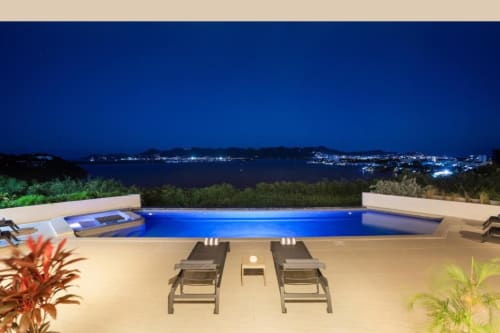 Villa de luxe avec piscine privée illuminée vue sur océan la nuit, deux transats sur terrasse, plantes tropicales, côtier