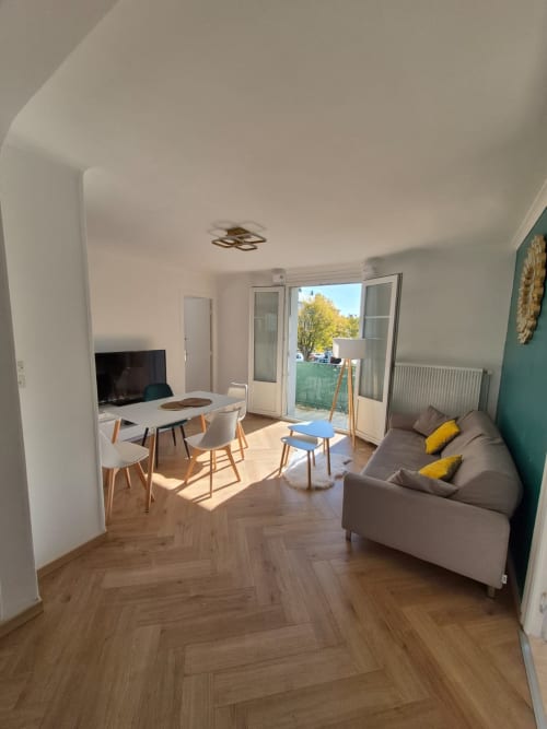Salon moderne d'appartement à Châteaudun avec canapé gris, téléviseur, coin repas et balcon donnant sur les jardins de la ville