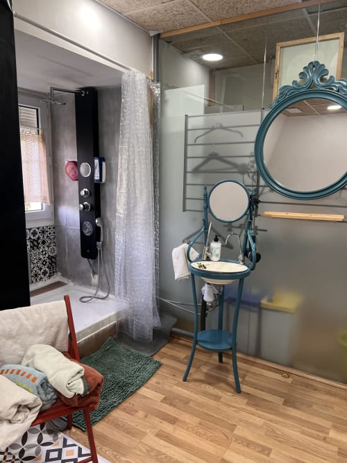 Baño moderno con ducha tipo walk-in, lavamanos tipo pedestal con espejo turquesa ornamentado, y piso de madera en los apartamentos Tessa